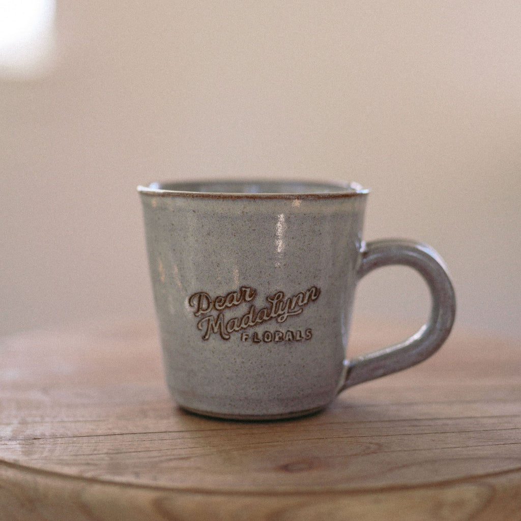 Gray ceramic mug with 'Dean Madalyn Florals' branding on a wooden surface.