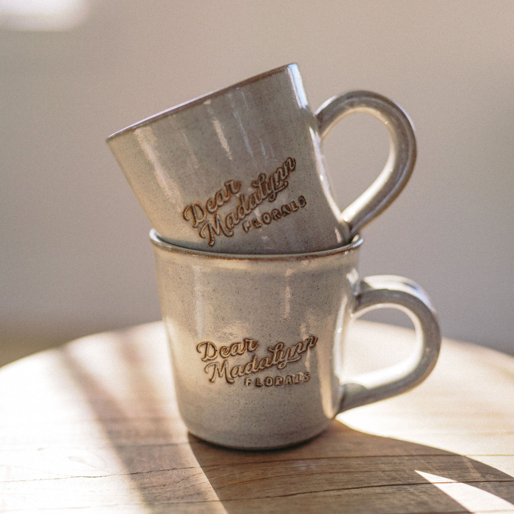 Two stacked metal mugs with 'Dear Madeline Florals' engraving on a wooden surface.